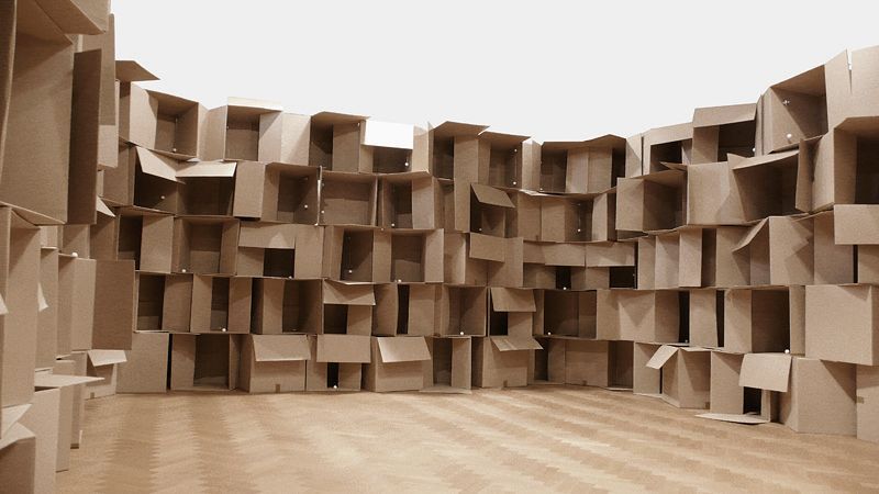 Room surrounded by tall stacks of open cardboard boxes, creating a maze-like interior on a wooden floor with a pale ceiling above.
