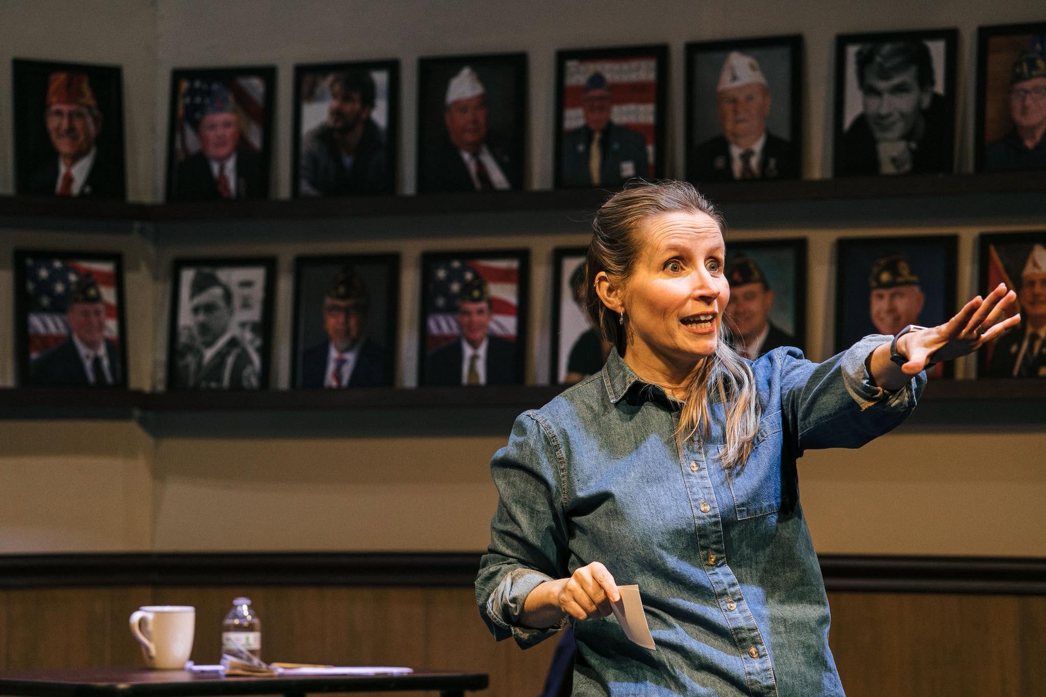 Woman in a denim shirt speaks, gesturing with her right hand as she stands at a table; framed portraits line the wall behind her.