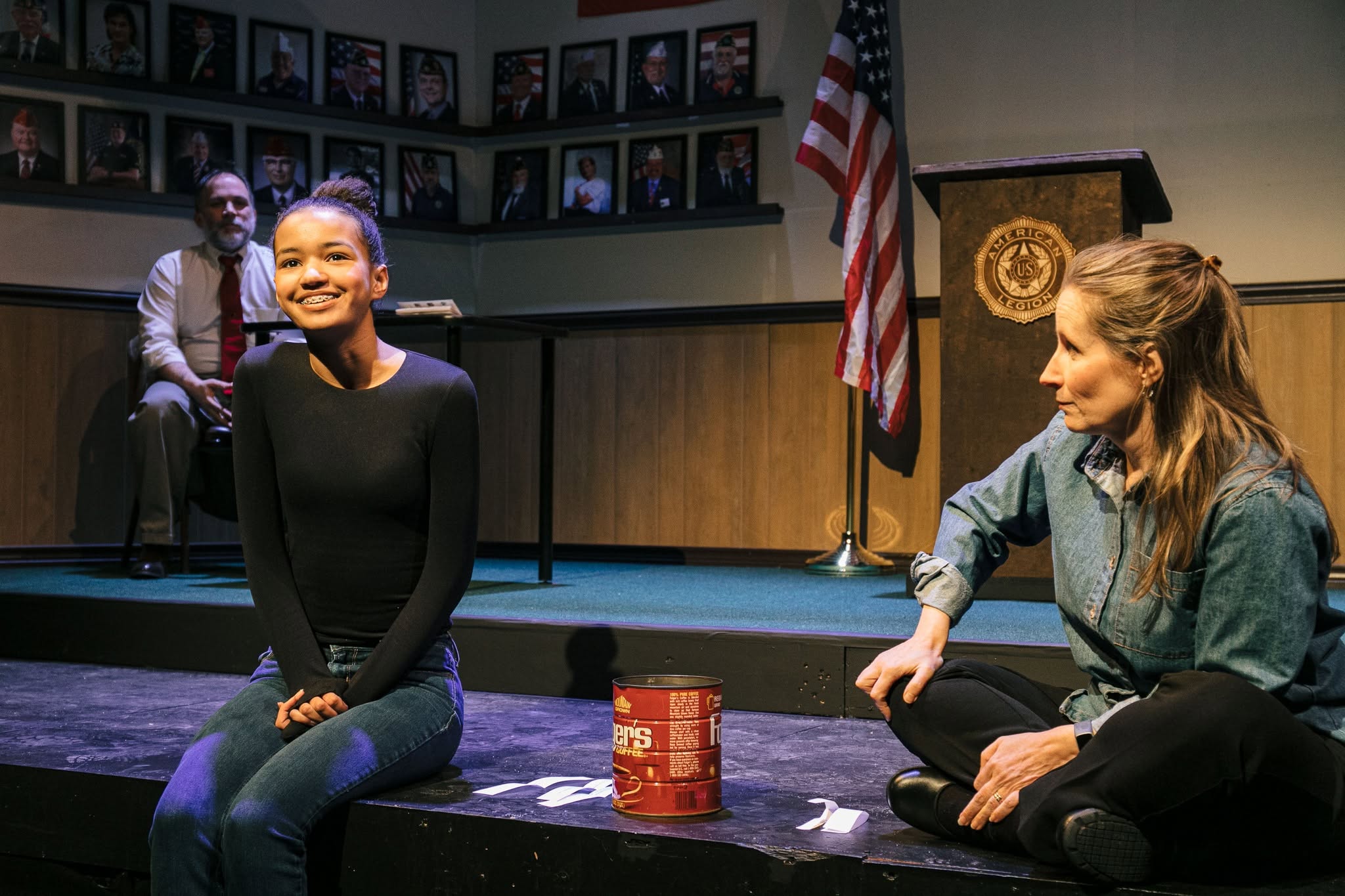 Two women sit on a stage during a community event; a man sits in the background, wall portraits line the room, and an American flag stands near a podium behind them.