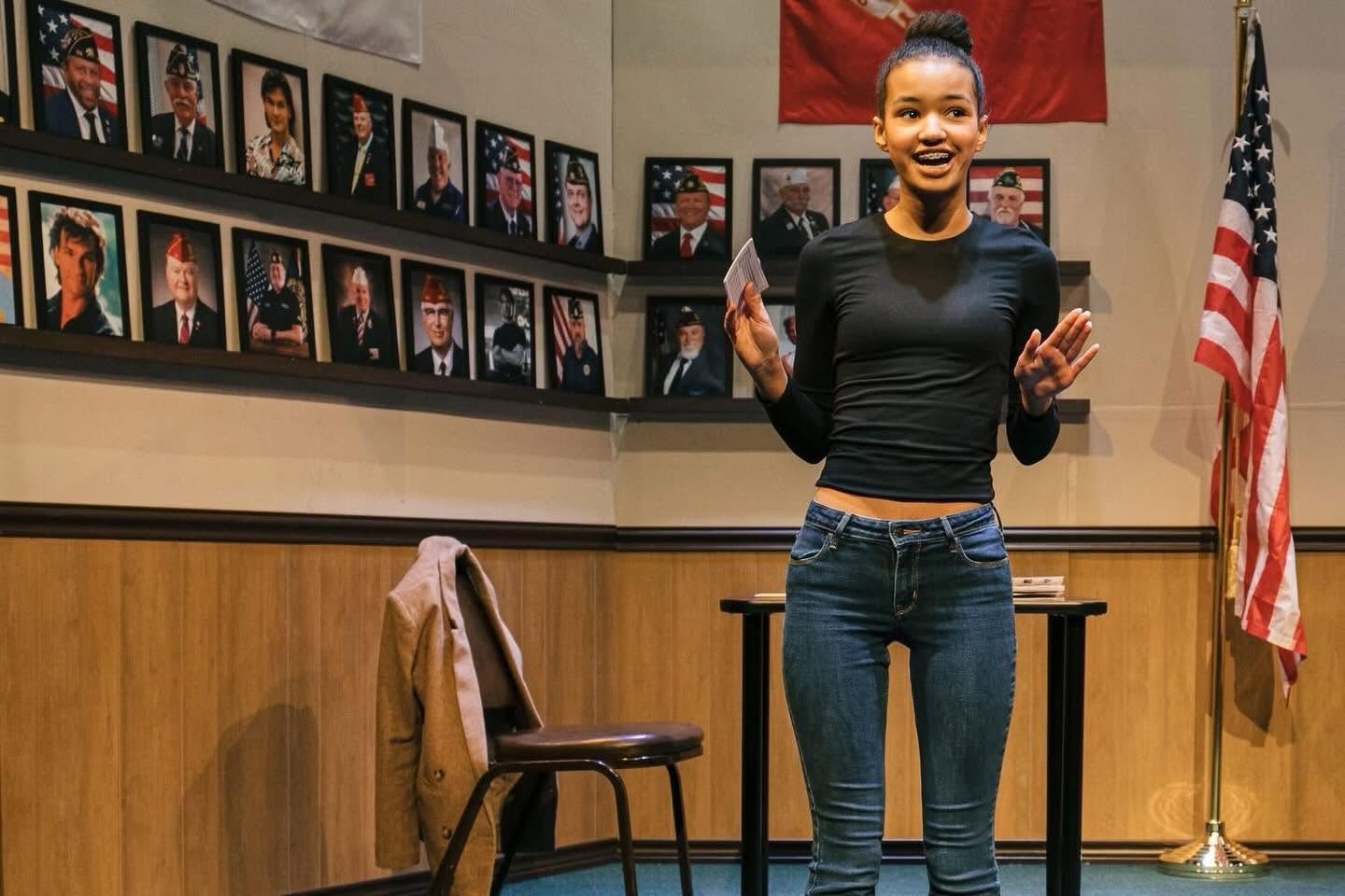 Young woman in a black long-sleeve top and jeans speaks at a podium in a room lined with framed portraits and an American flag nearby.