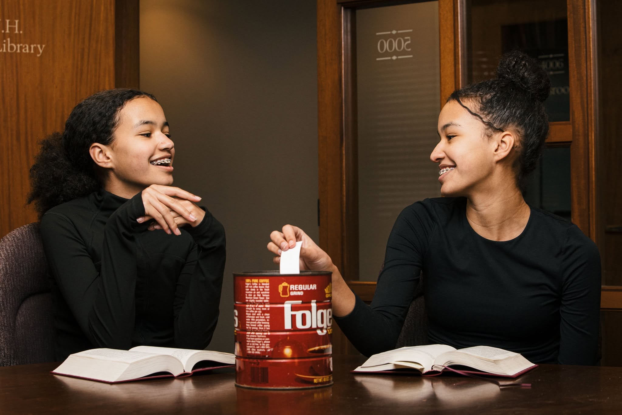Two girls in black shirts sit at a table with open books, smiling at each other; a large red Folgers coffee can sits between them in a library-like setting.