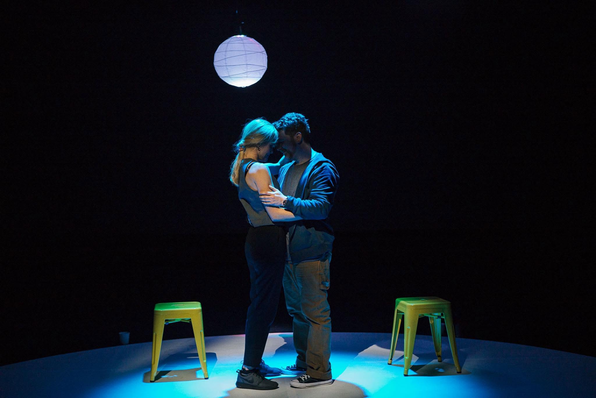 Two people share a close, intimate moment on a lit circular stage under a hanging paper lantern, surrounded by colorful stools in blue light.