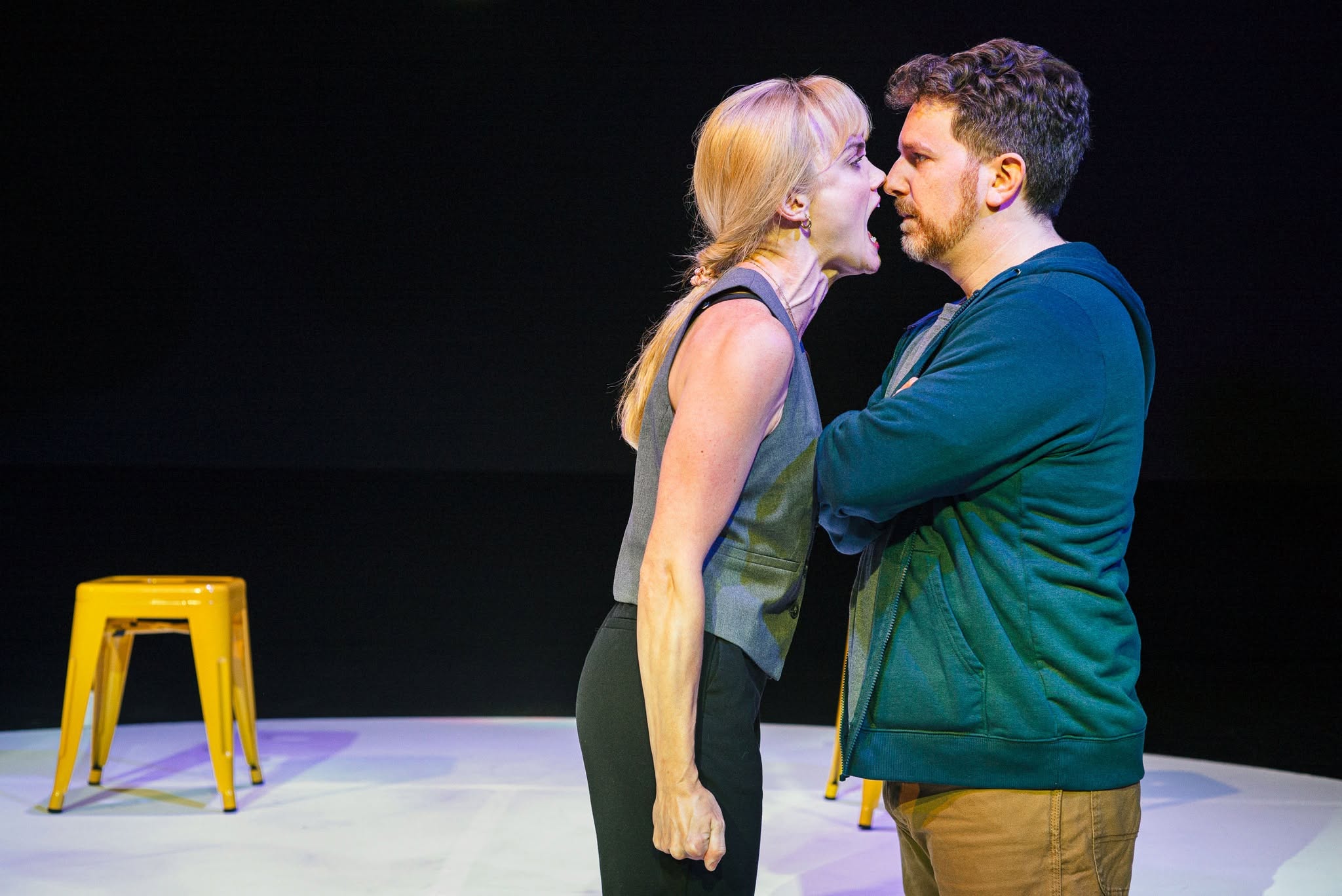 Two adults on a stage lean toward each other with mouths open, as if arguing, with yellow stools visible on the left.