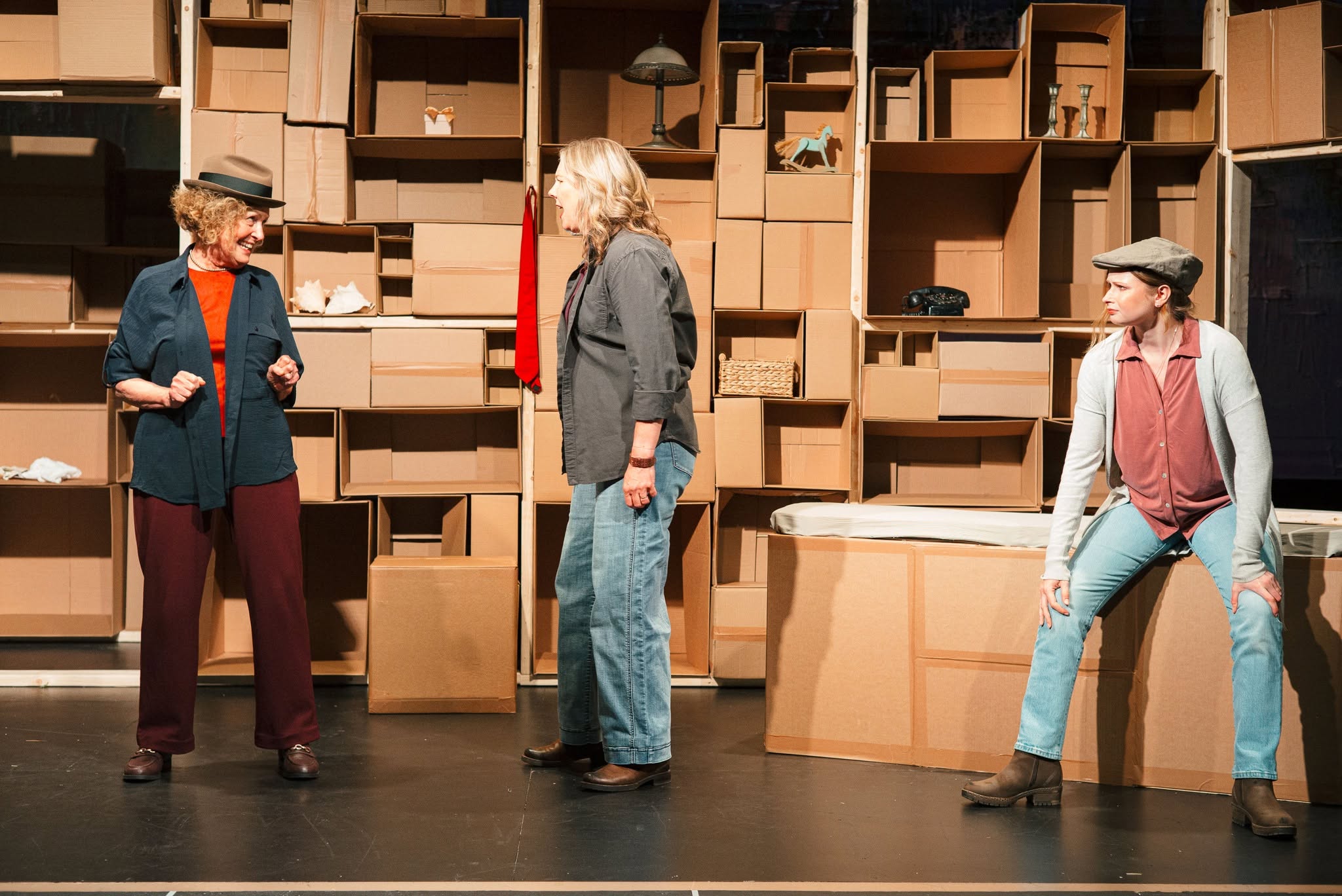 Three performers on a stage beside a wall of stacked cardboard boxes, smiling and talking together.