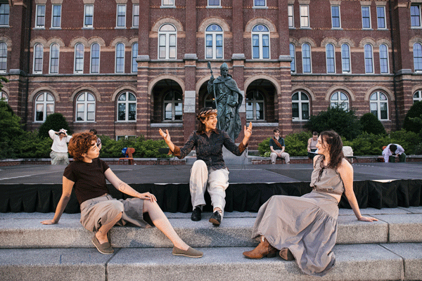 Three people sit on stone steps in front of a bronze statue and a red-brick building, chatting and gesturing.