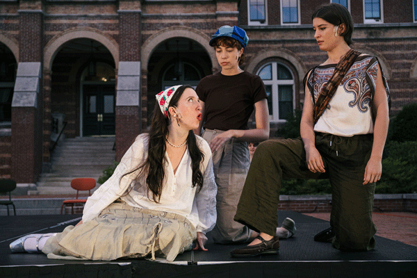Three women perform a dramatic scene on an outdoor stage in front of a brick building: one seated on the ground in a white blouse and headscarf, another kneeling with a blue cap, and a third standing with a patterned sleeveless top and olive-green pants.
