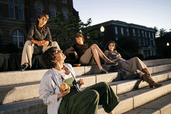 Five people sit on wide concrete steps outside a brick building, relaxing and chatting in the late afternoon light. A man in green pants holds an apple in the foreground.