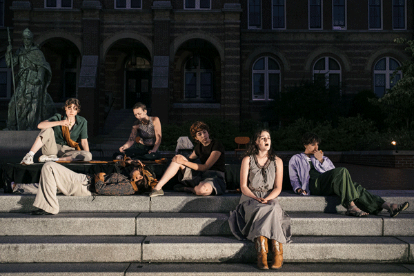 Six young people sit on wide stone steps in front of a brick building at dusk; some are talking, one reclines, and a woman in boots sits near the center, creating a relaxed, social scene.