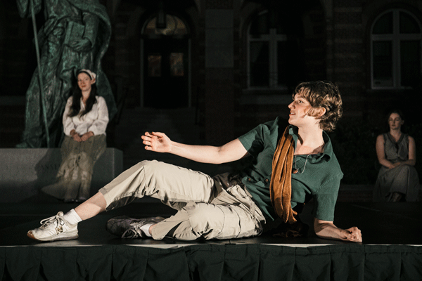 Young man in a green shirt and beige pants lounges on a theater stage, with a statue and two seated people in the background.