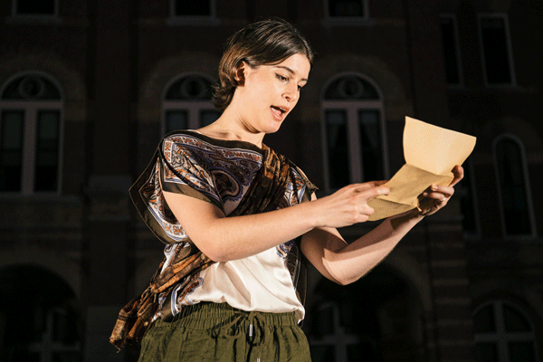 Woman on stage, reading a letter from a yellowed envelope, wearing a patterned shawl and green pants at night outside.